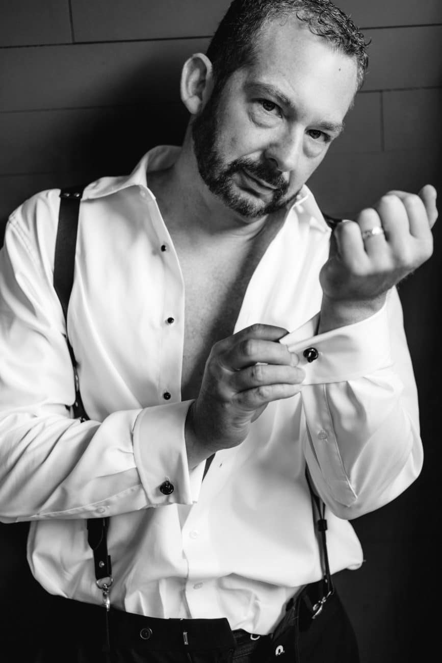 Black and white photo of man in white shirt and suspenders, cuffing sleeves, looking into camera, men's boudoir