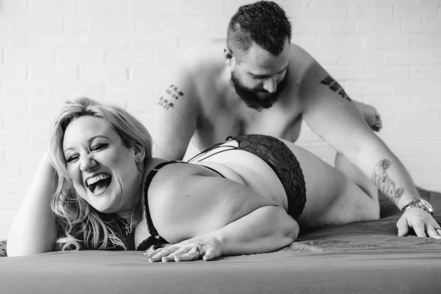 Black and white couples boudoir photo of woman on her belly laughing while her partner plays drums on her booty