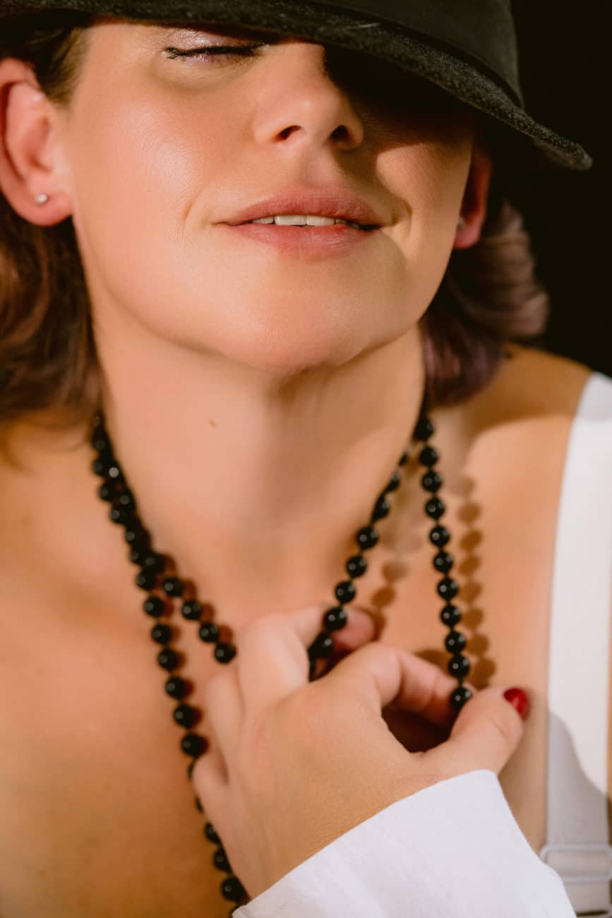 Close up boudoir photo of a woman in a hat, with a slight smile, grabbing her black pearl necklace