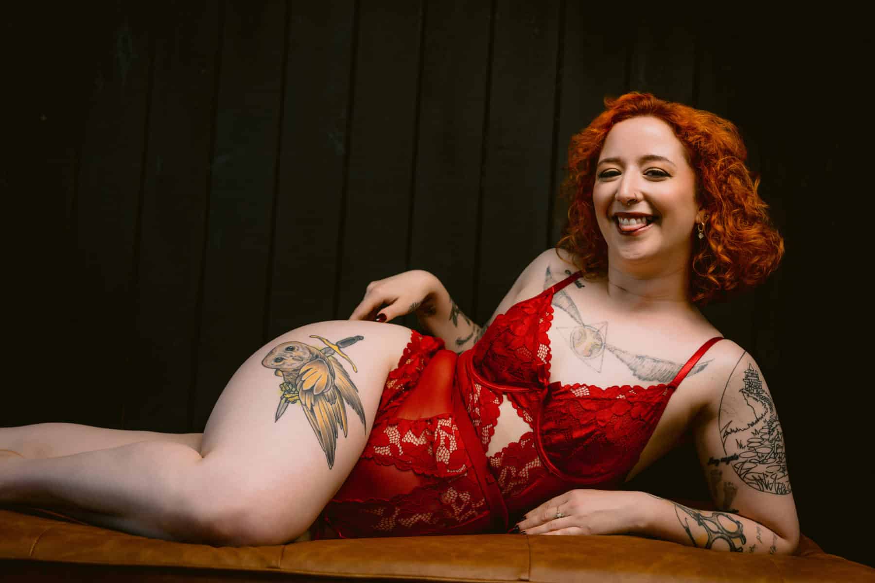 Boudoir photo of a woman in a red lace bodysuit with bright orange hair, laying on her side with a cheeky, tongue-out smile. 