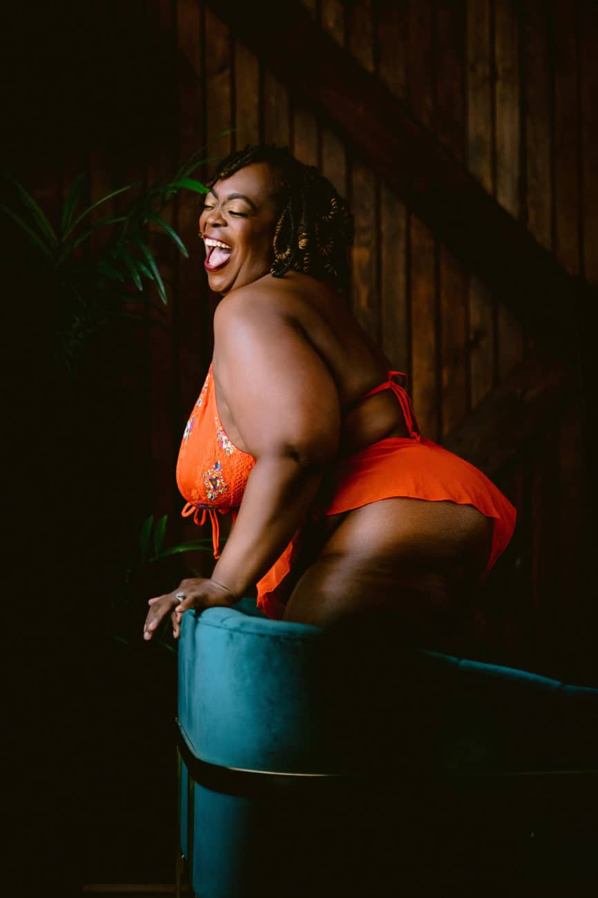 Plus size black woman boudoir photo in a Savage Fenty, bright orange bodysuit laughing