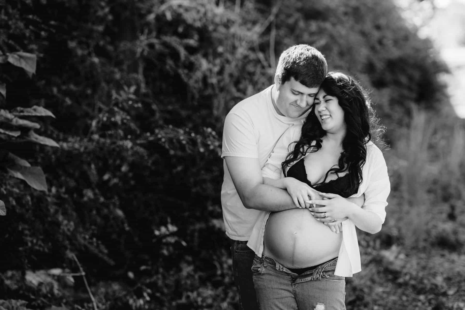 Black and white photo of dad to be holding pregnant wife, with open white button down and bare belly.