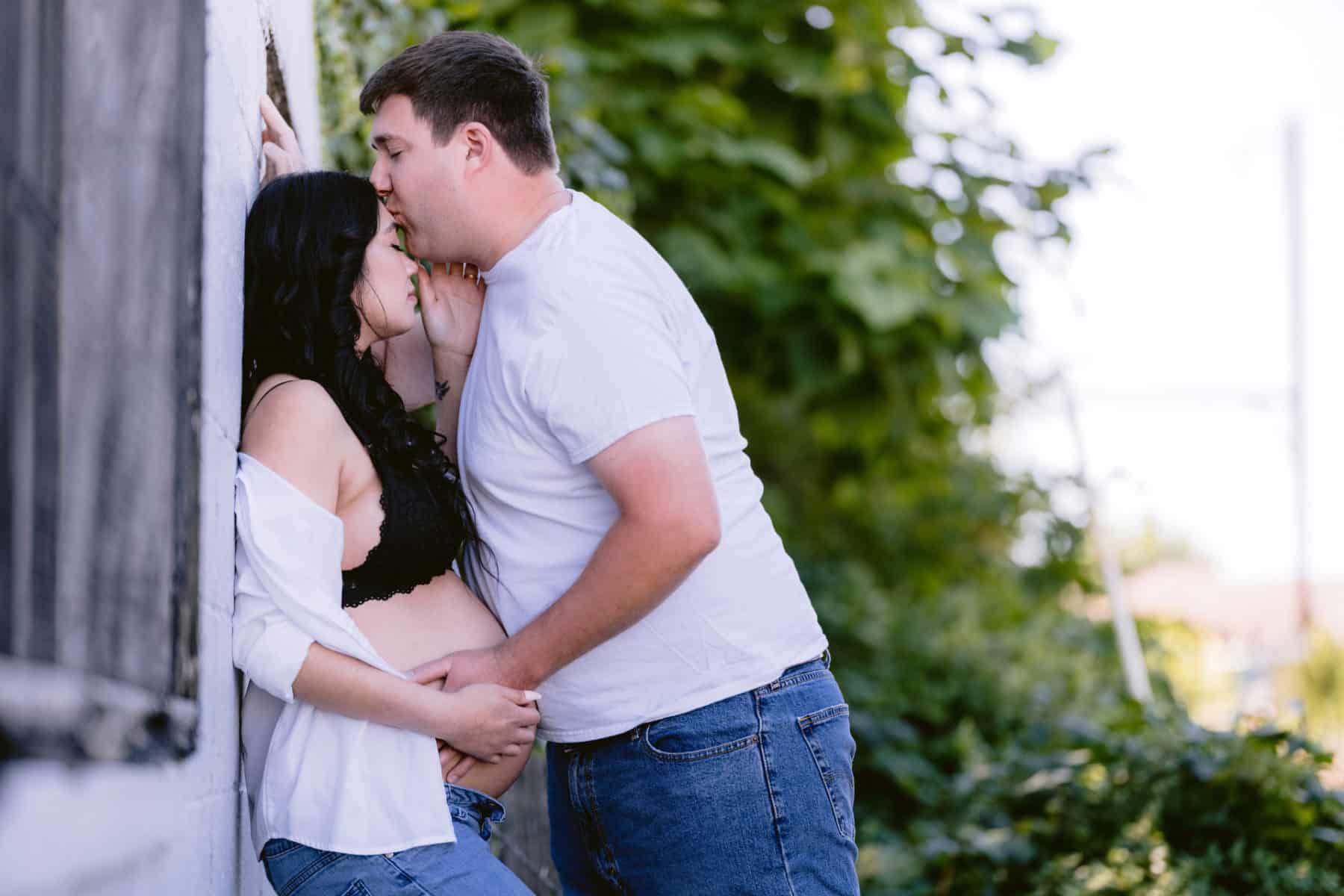 Outside photo of expecting parents, dad kissing mom's forehead, while they both hold her bare pregnant belly.