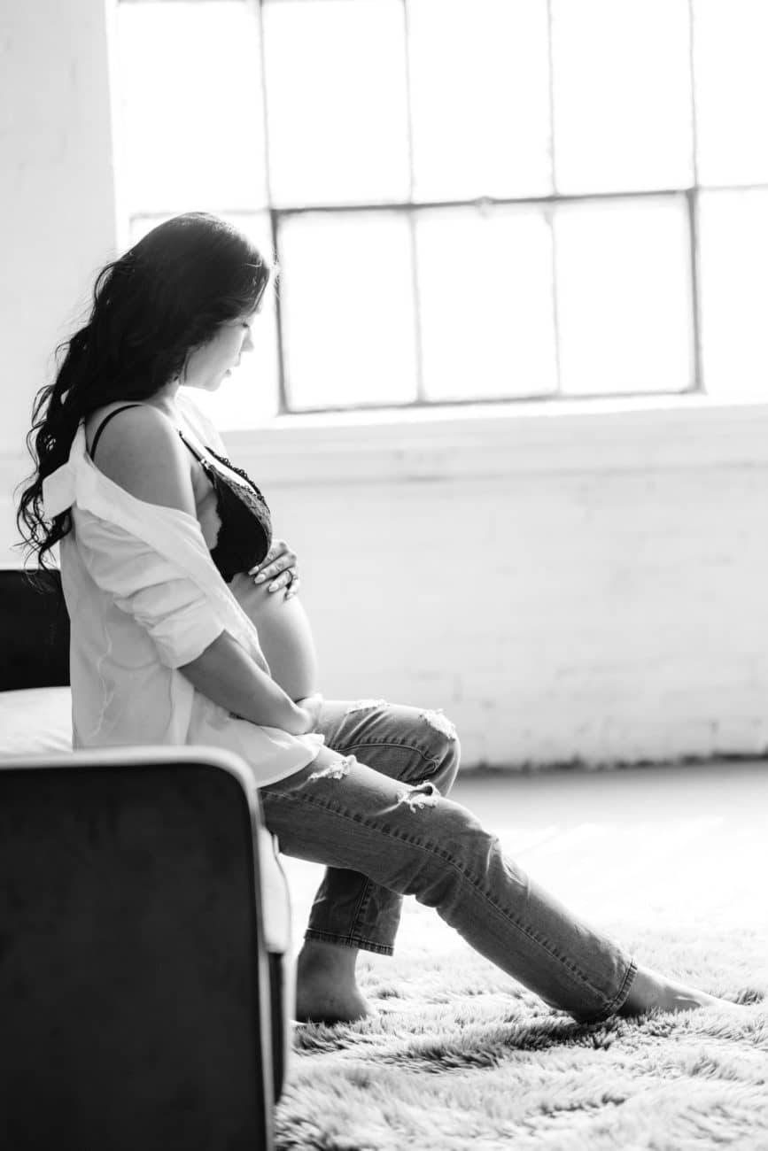 Black and white pregnant woman in industrial studio, holding bare belly, with jeans and white opened button down.