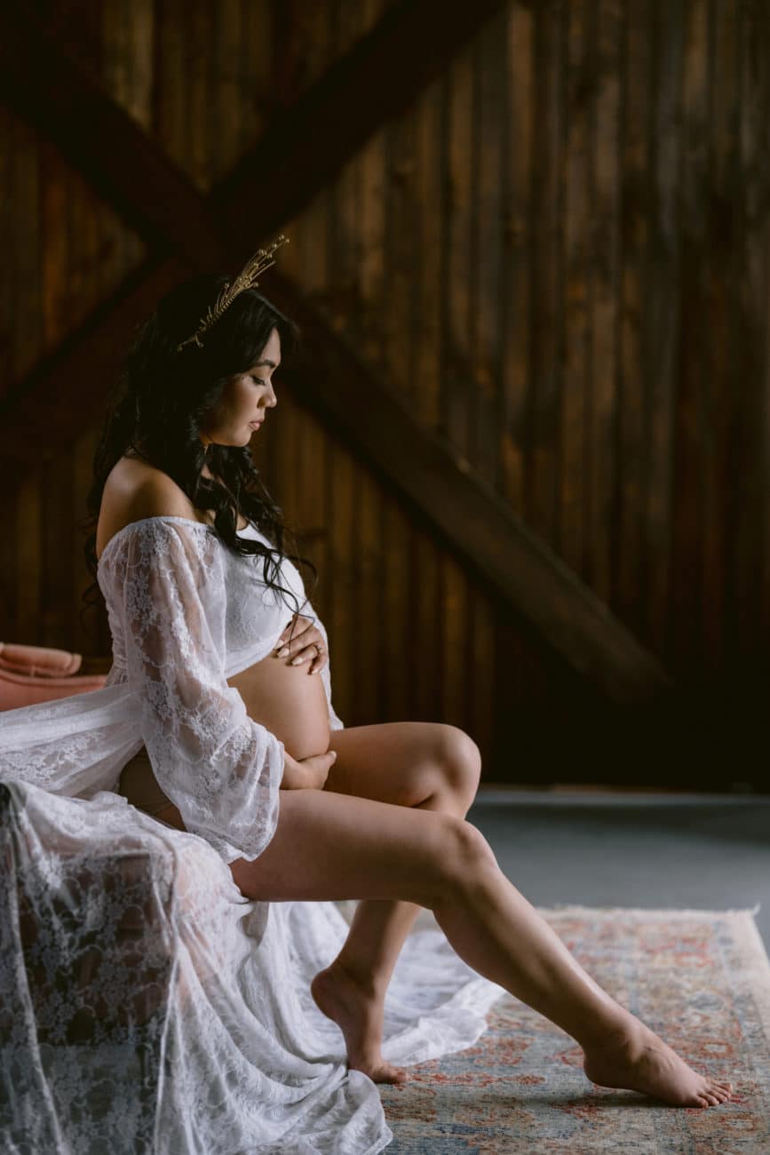 Pregnant mom to be holding bare belly, wearing white lace maternity robe and gold sun crown.