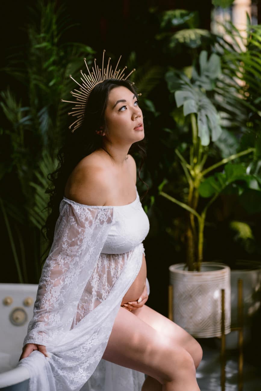Goddess-like pregnant mom in white lace maternity robe, exposed baby belly, and golden sun crown.