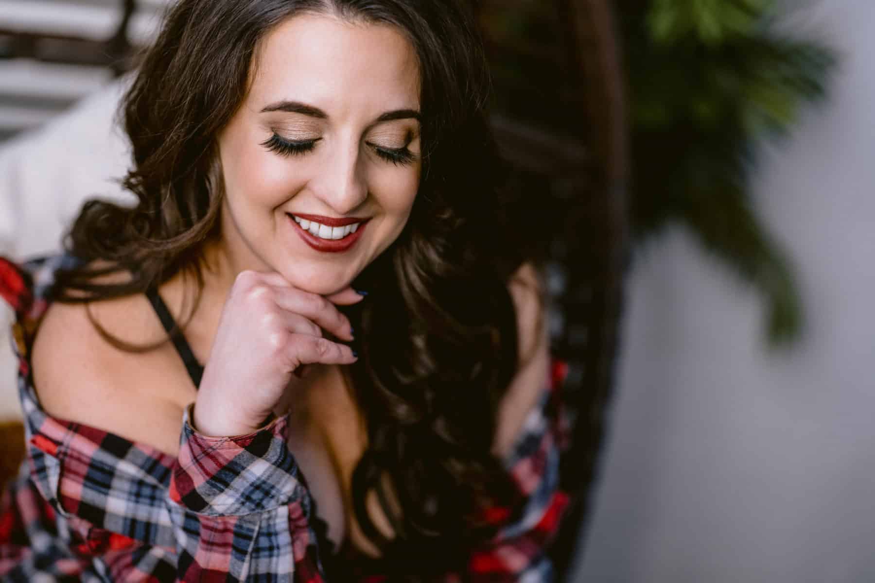 Photo of woman glancing down and smiling with her hand below her chin, in fiance's plaid shirt falling off shoulders